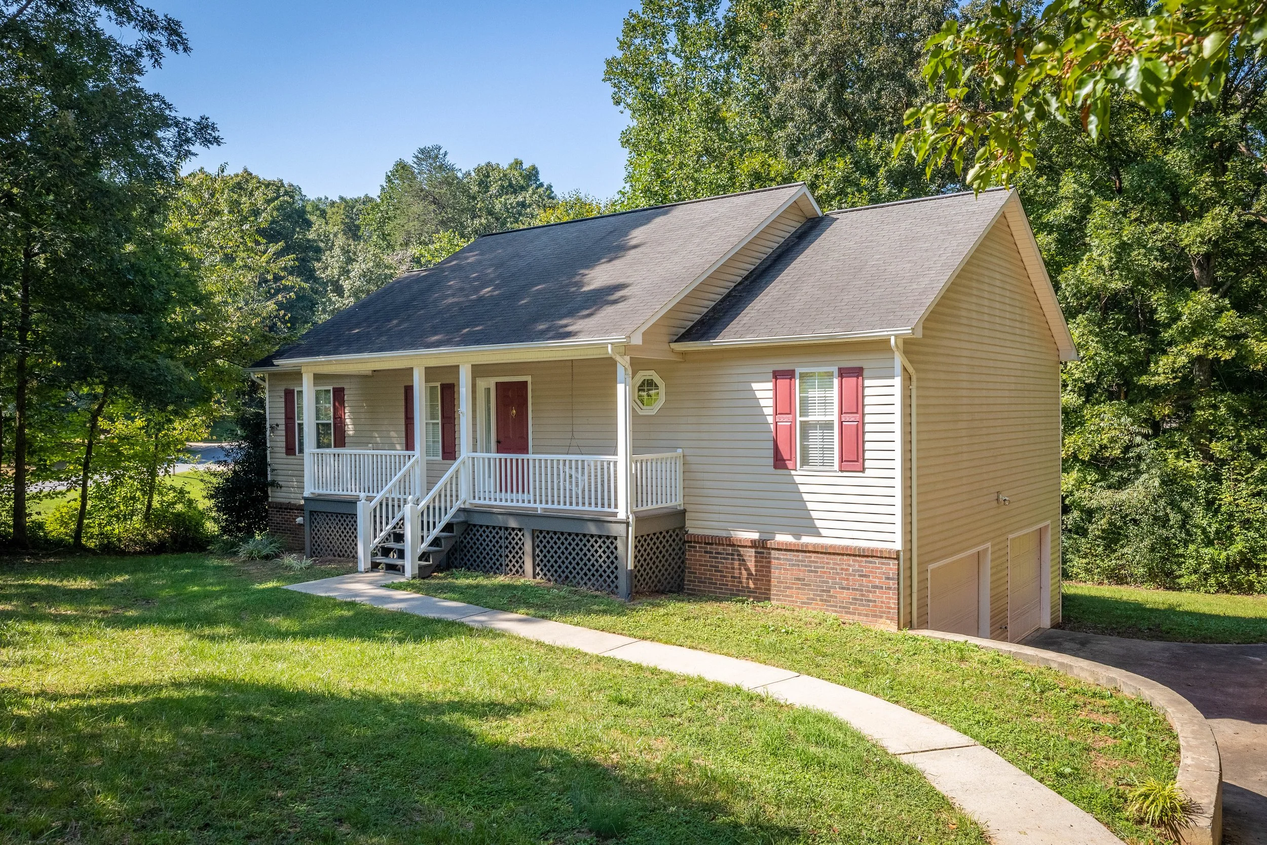 A beige house with red shutters, a front porch with white railings, and a two-car garage at the basement level, surrounded by green grass and trees. Great Rental property.