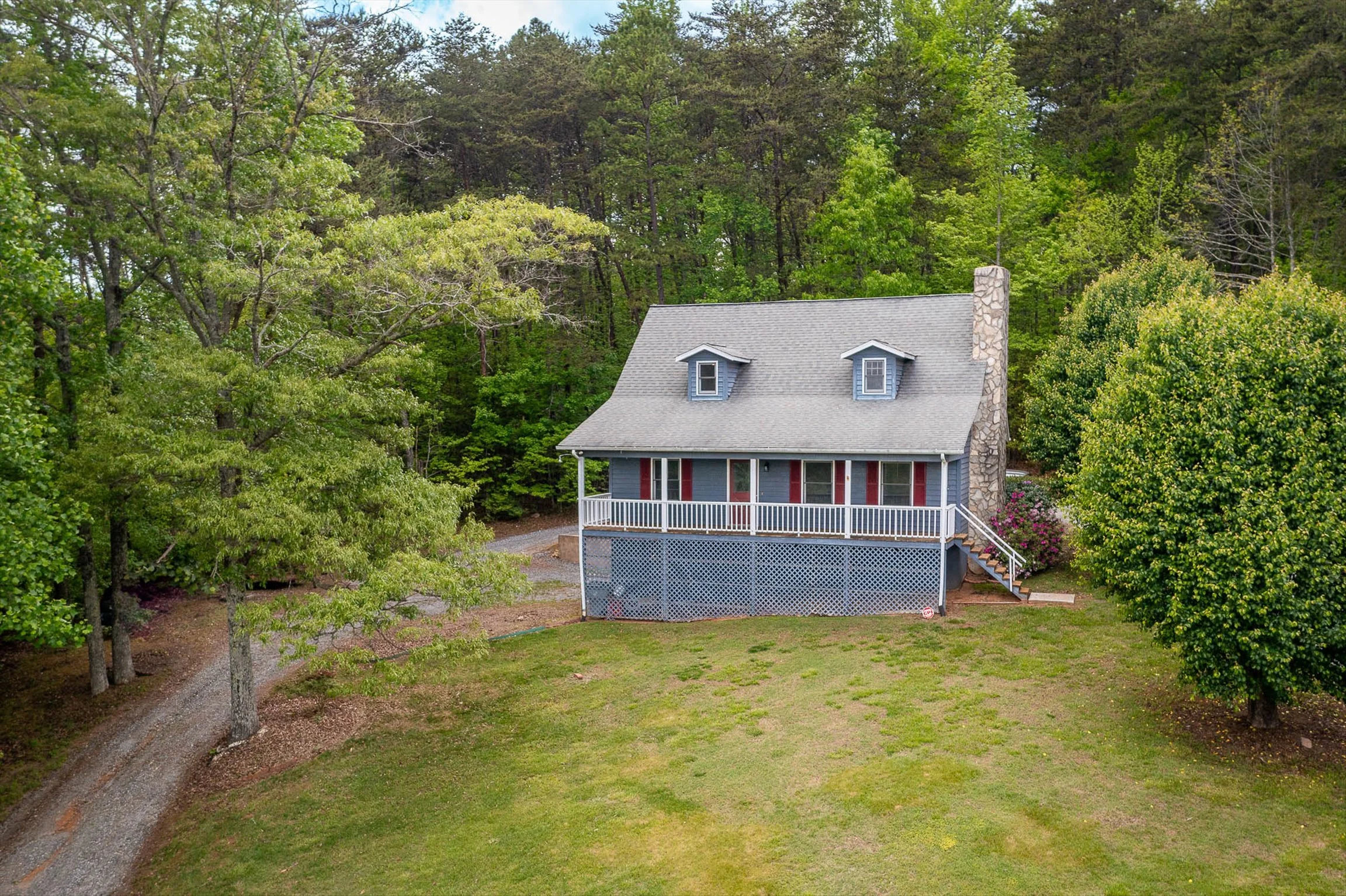 A blue house with a gray roof and red shutters, surrounded by trees and a grassy yard, with a gravel driveway on the left. Beautiful 3 story home that has a lot of rental potential.