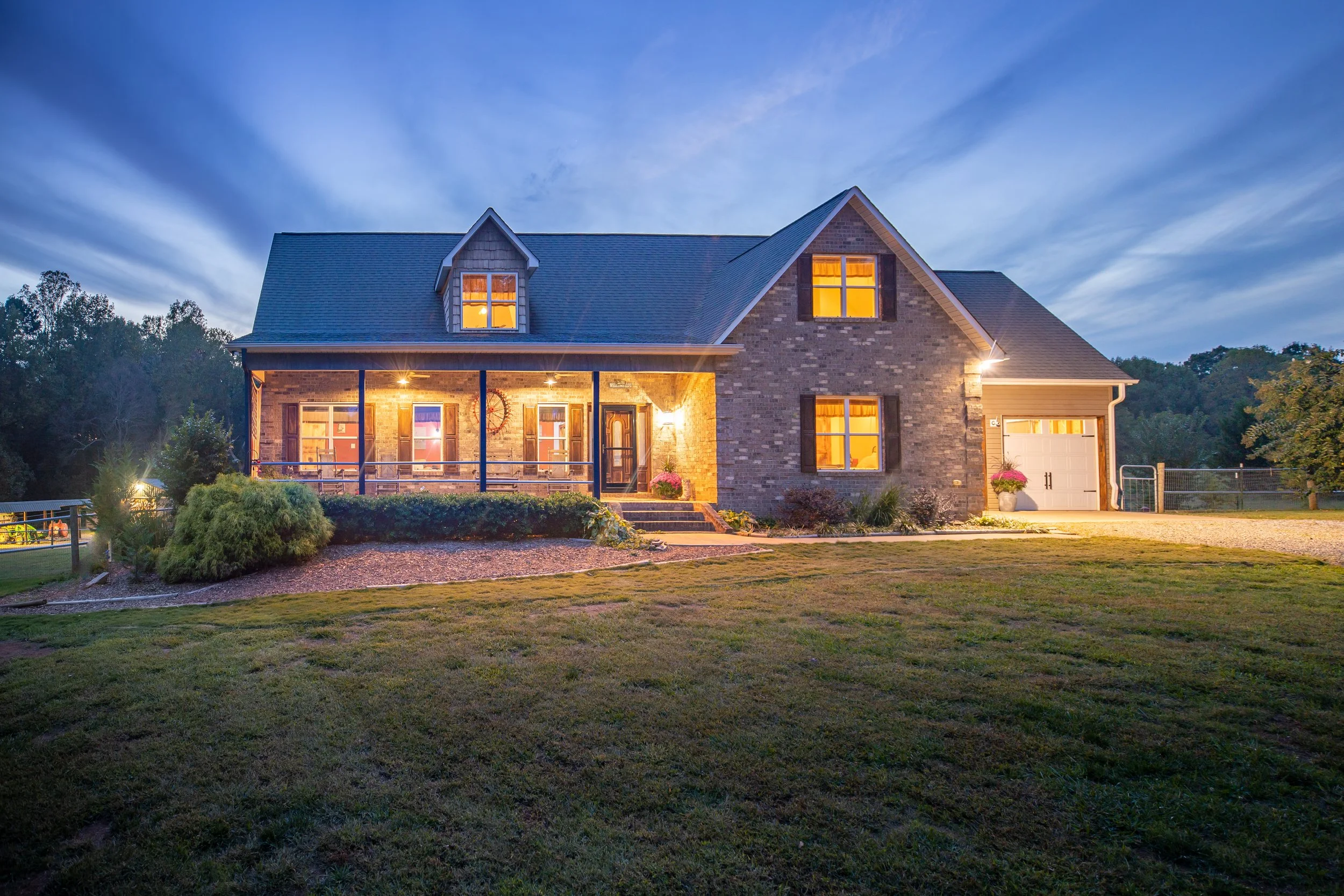A large brick house with warm lit windows and porch lights, a well-maintained lawn, and trees in the background at dusk.