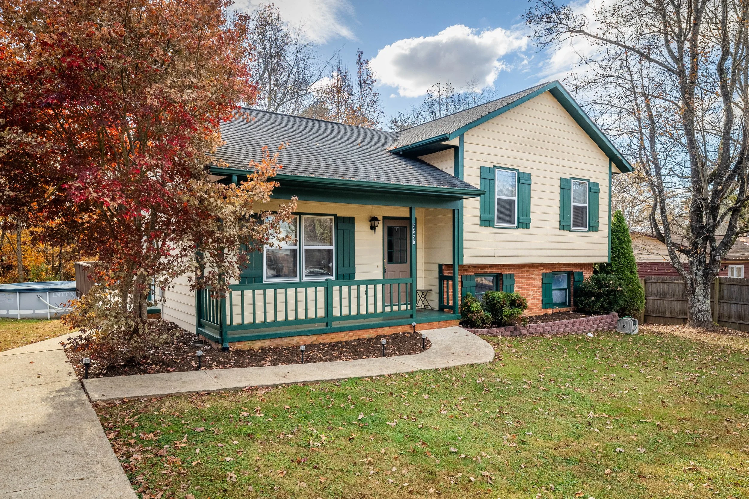 A two-story house with yellow siding, green shutters, and a chimney, surrounded by a lawn and trees with fall foliage. Great starter home for people to get into the real estate market.