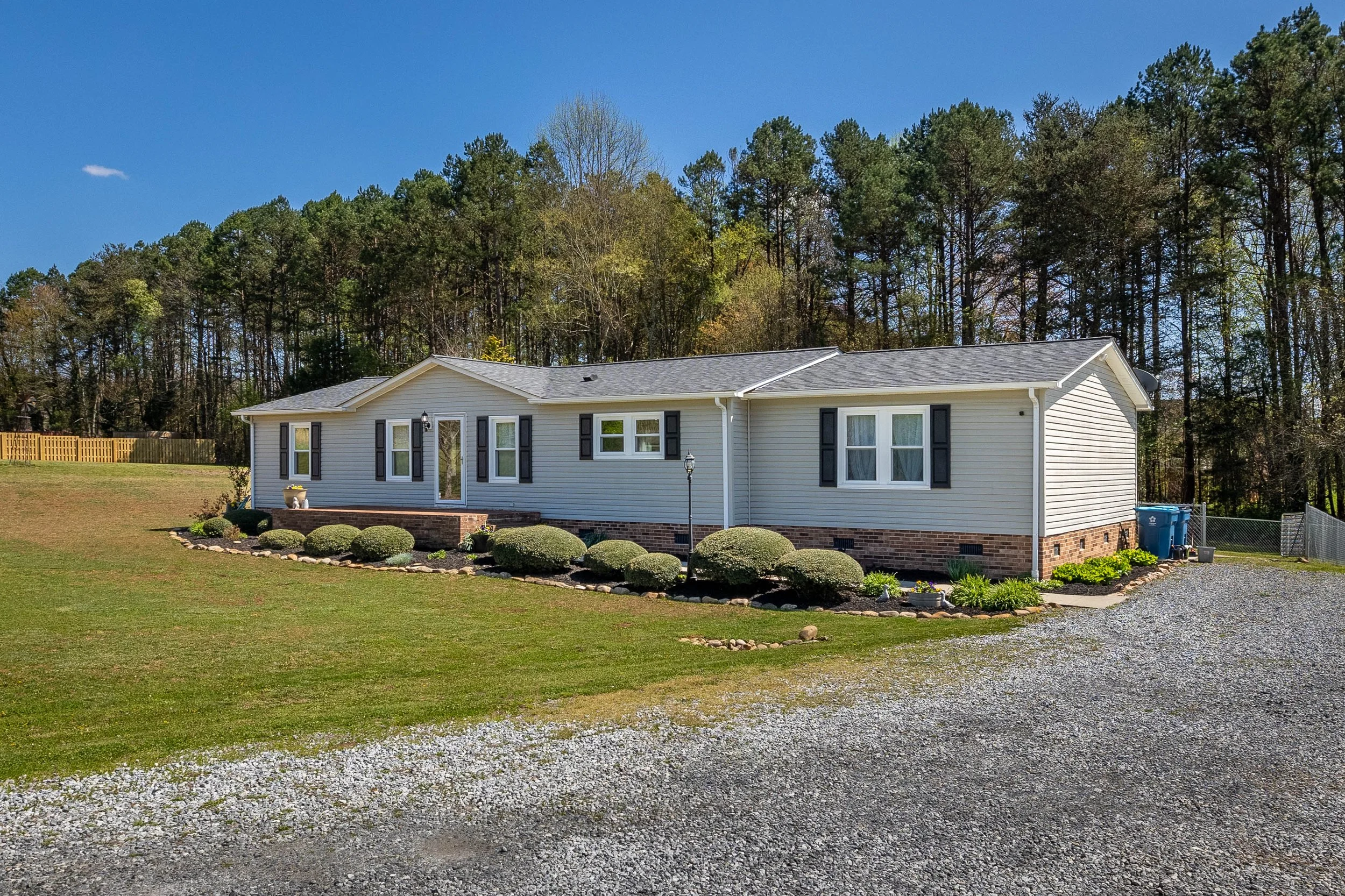 A white manufactured home with black shutters, situated on a grassy yard with landscaped bushes, a small brick porch, and a gravel driveway. There are trees in the background under a clear blue sky. Rural living for tenants to make them at home.
