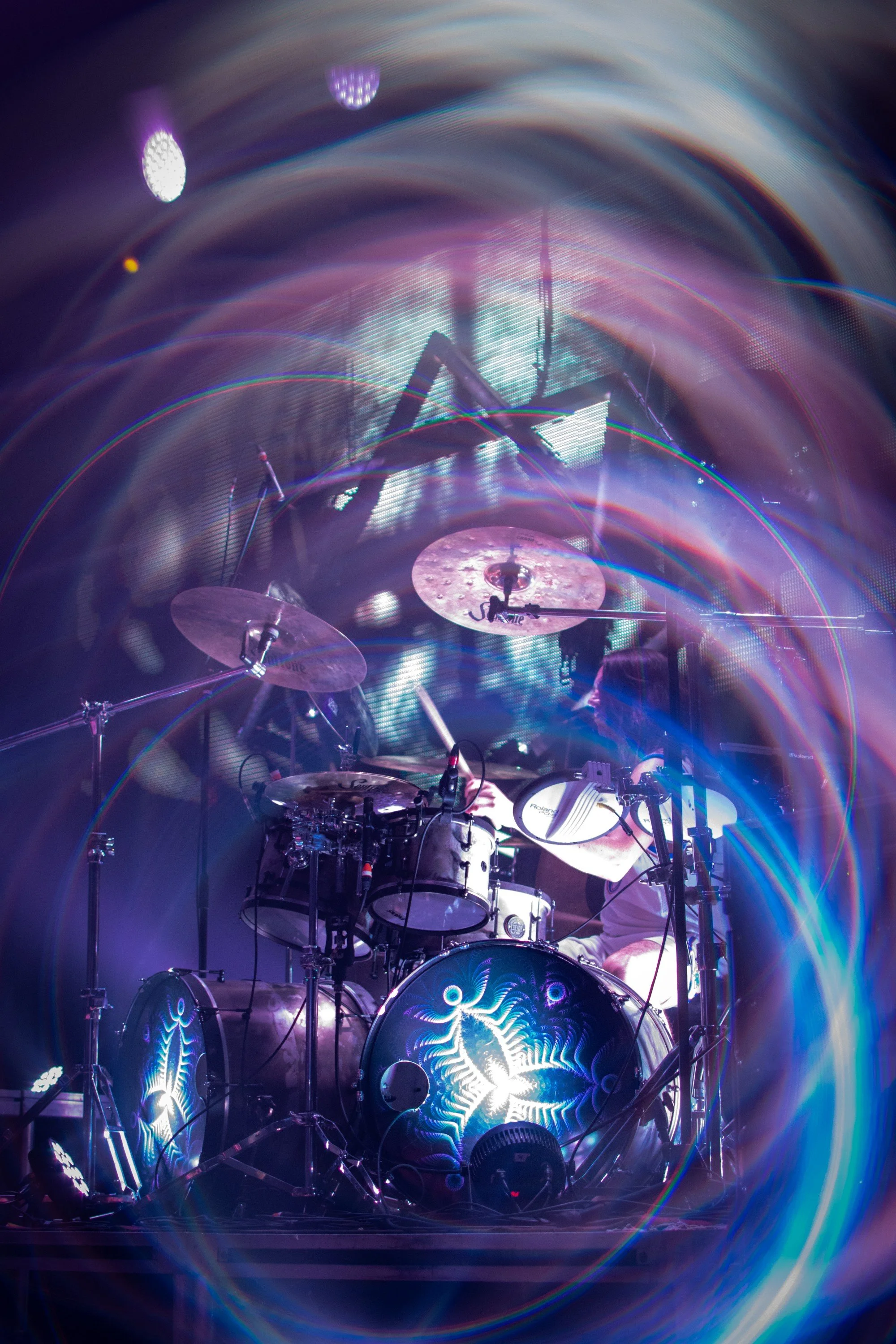 A person playing a drum set on stage with colorful lights and laser effects creating a psychedelic atmosphere.