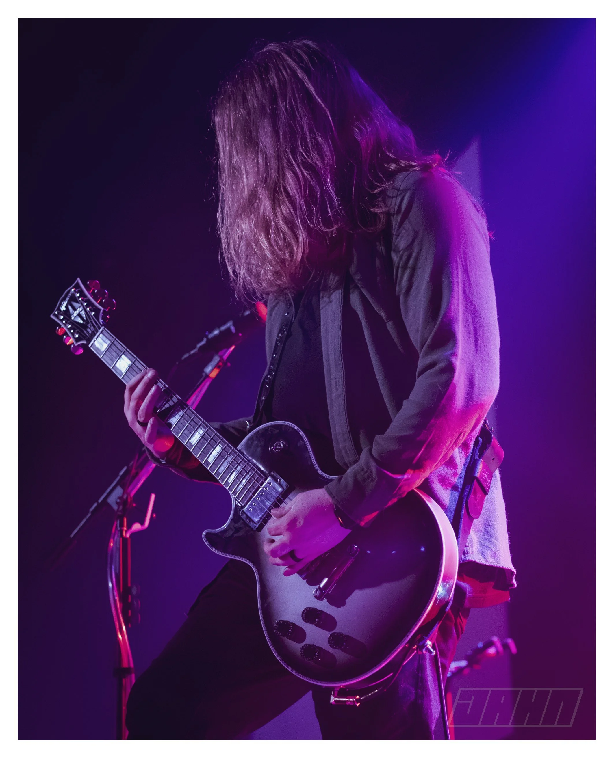 Musician playing an electric guitar on stage illuminated with purple and pink lighting.