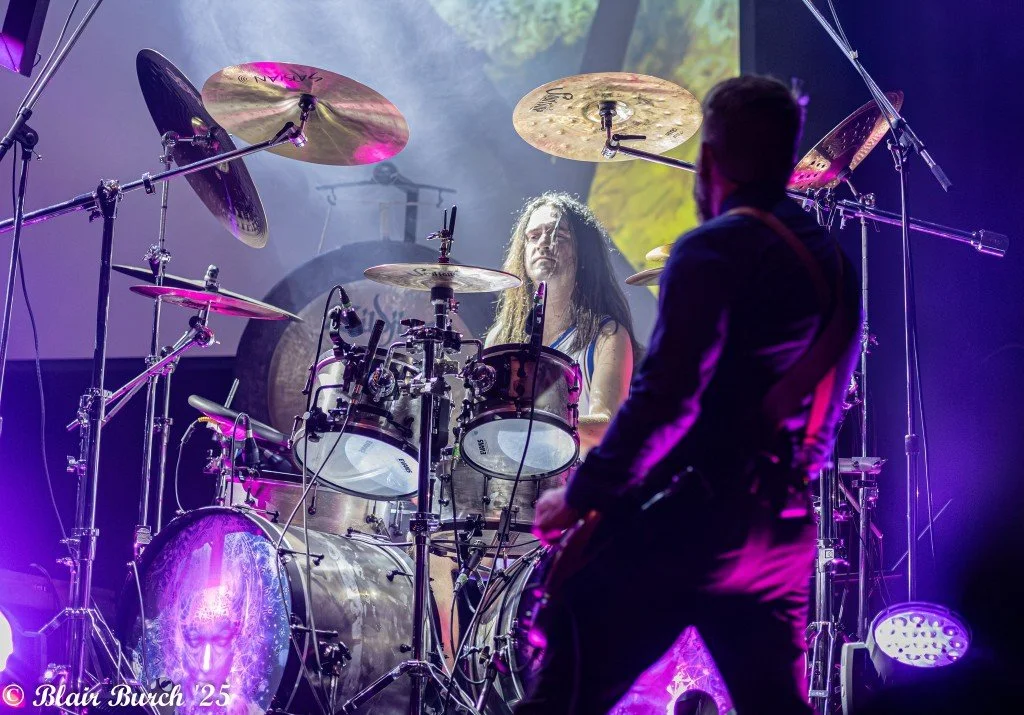 A female drummer with long hair playing a drum set on stage, seen from the front, with a male guitarist in the foreground.