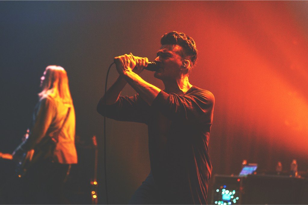 Musician singing into a microphone on stage with warm orange lighting, with a female band member in the background.