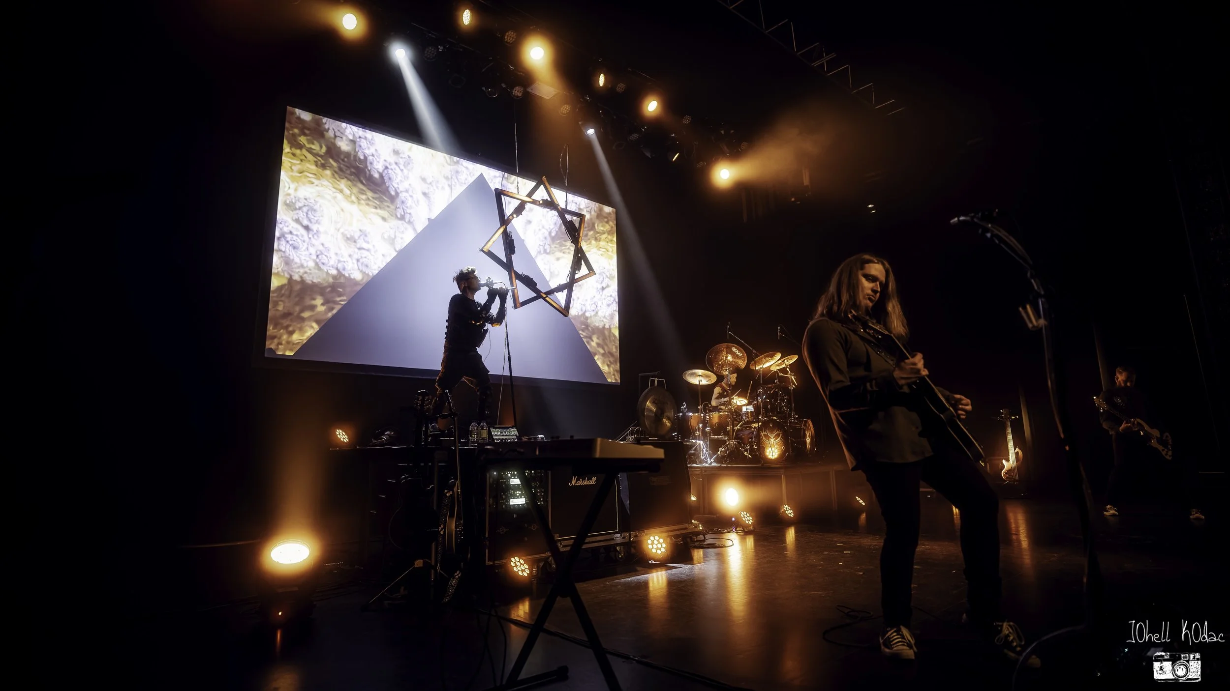 A band performs on stage with lighting and visual effects, featuring musicians playing instruments such as guitar, drums, and wind instrument, with a large screen in the background displaying abstract visuals.