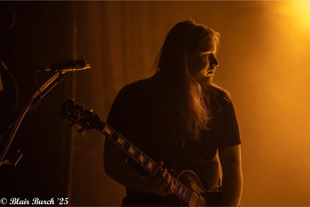 A woman with long hair holding an electric guitar in a dimly lit setting with warm orange lighting.