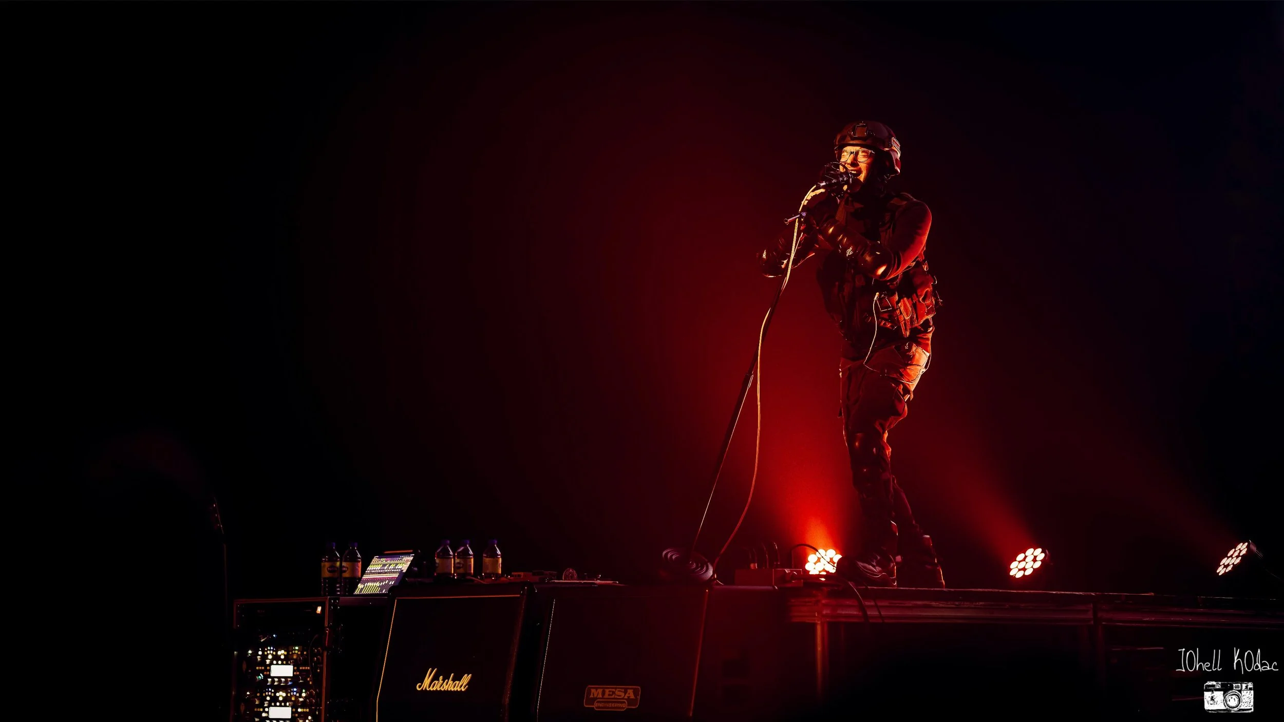 A person dressed in military-style clothing, with a helmet and gloves, singing into a microphone on stage with red stage lights highlighting them.
