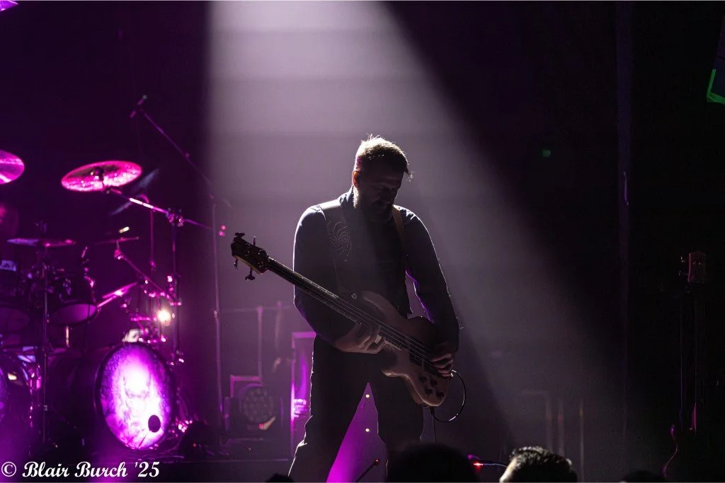 A musician playing an electric guitar on stage, backlit with purple and white lighting, with a drum set visible in the background.