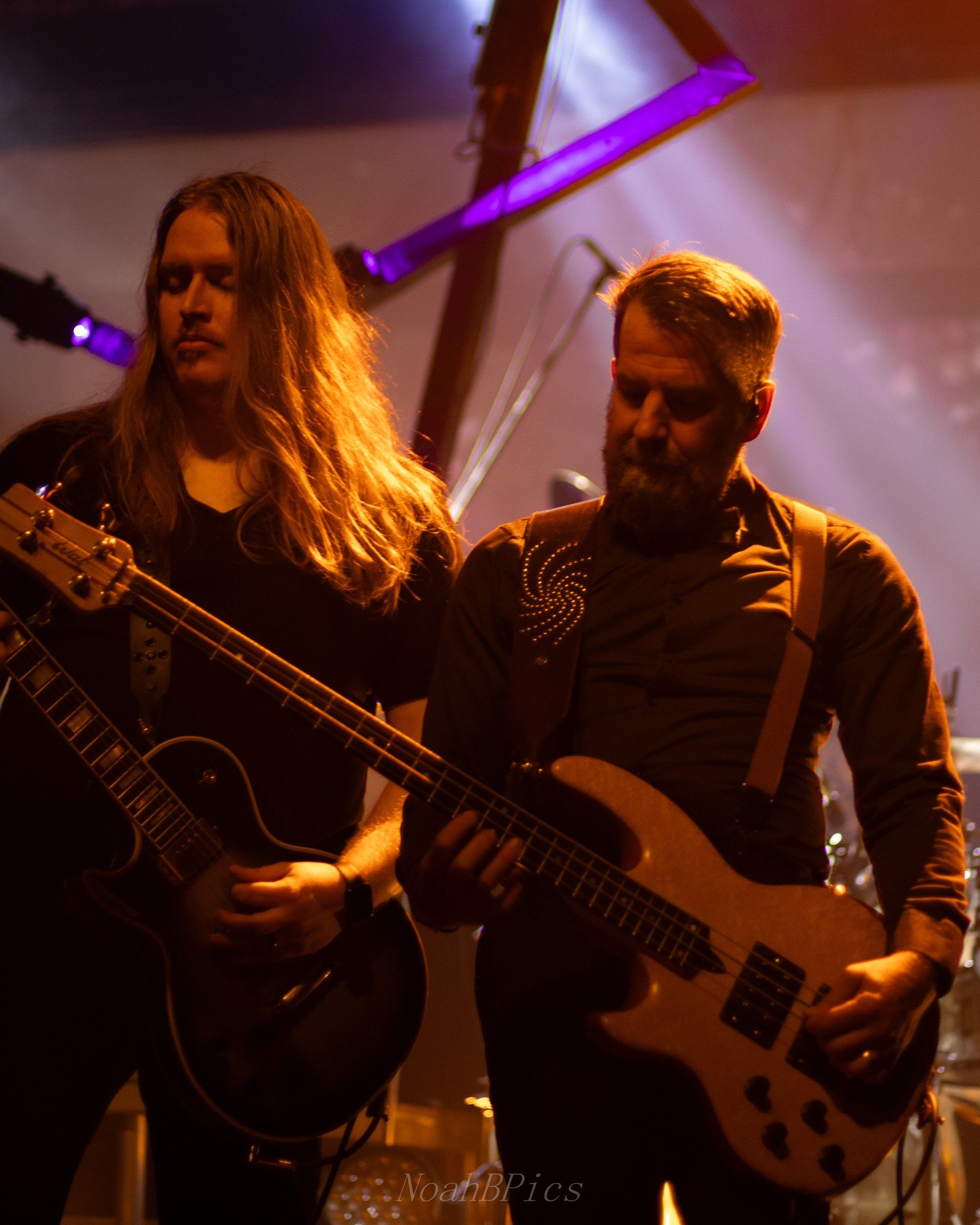 Two musicians, a woman and a man, playing guitars on stage under purple and orange lighting, with stage equipment in the background.