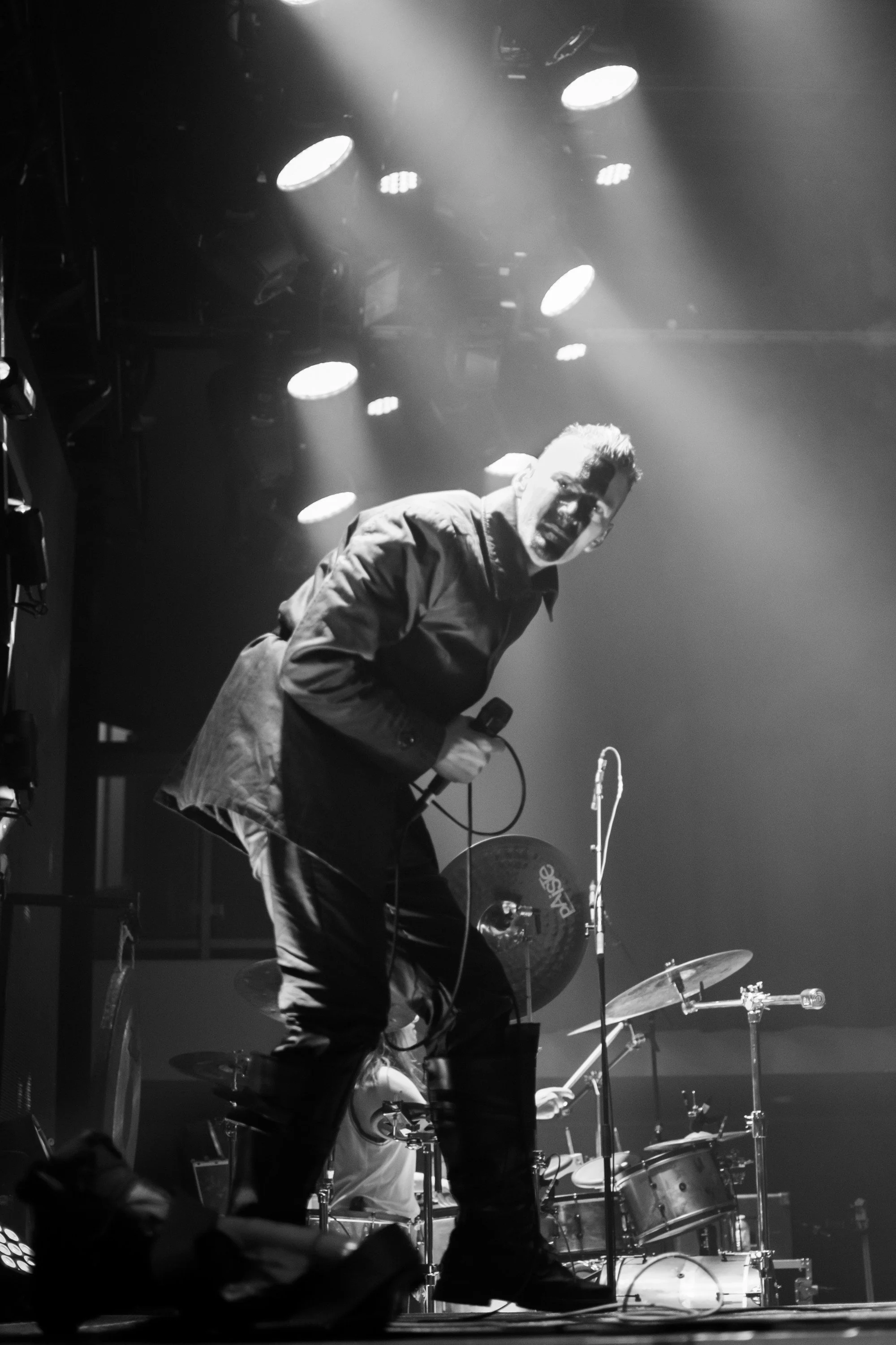 A man performing on stage, holding a microphone, with drums and stage lights in the background, in black and white.