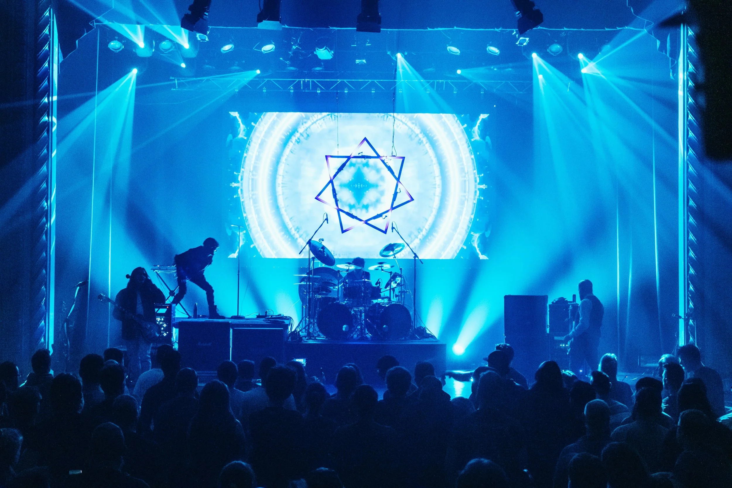 Concert stage with blue lighting, a band performing, and a large digital screen displaying a geometric star pattern in the background.