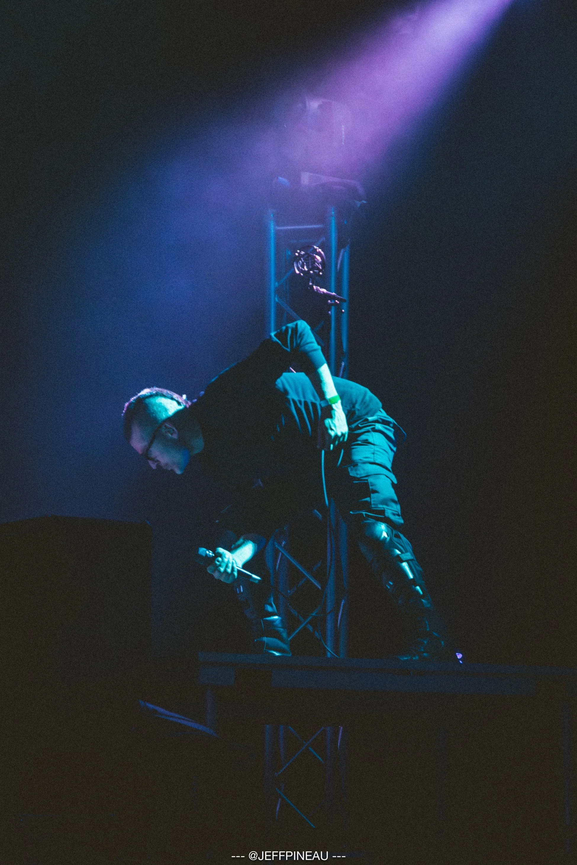 A man is on an elevated platform with a microphone in hand, illuminated by colorful stage lights, likely speaking or singing during a performance.