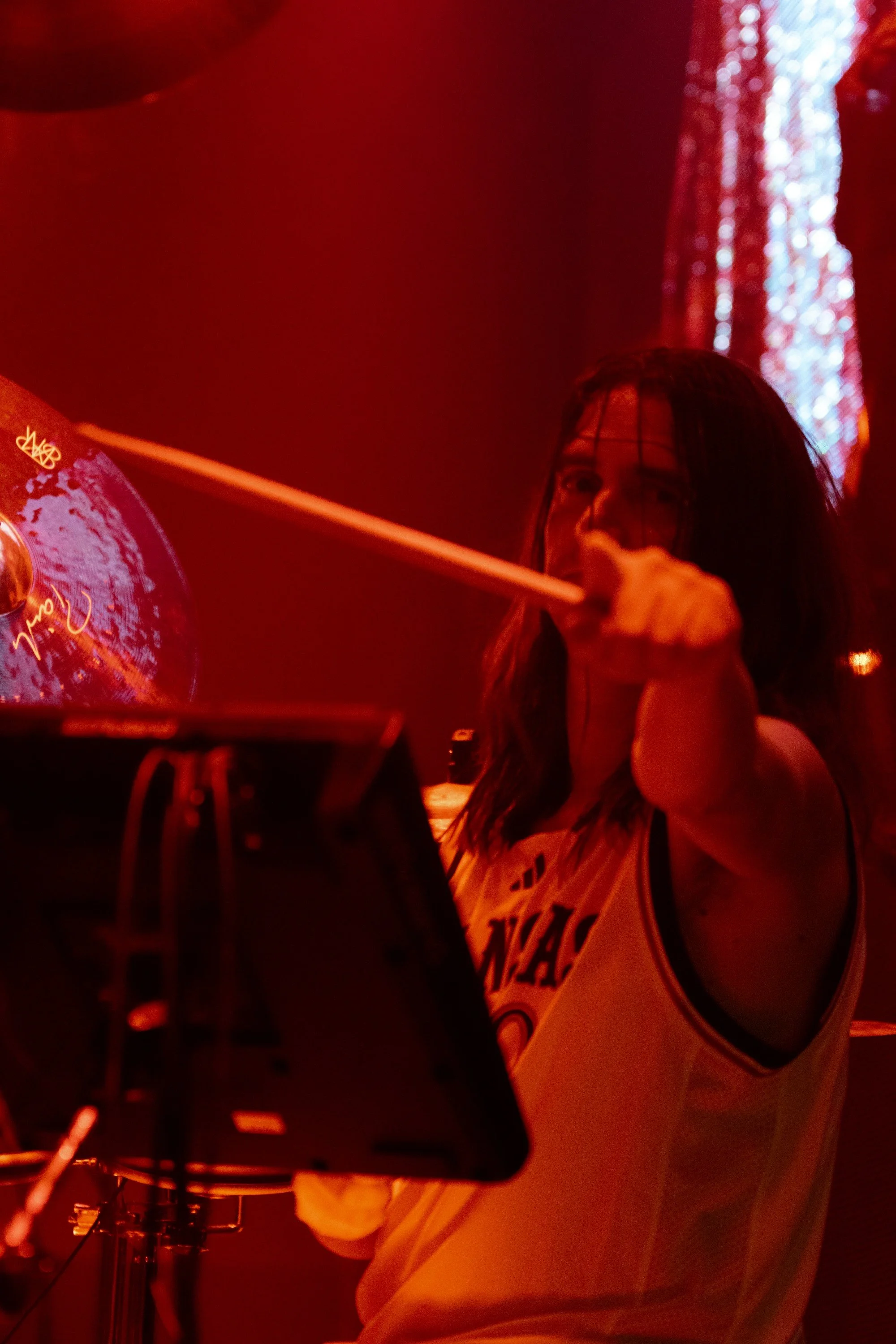 A woman with dark hair is playing drums with drumsticks in a dimly lit room with red lighting.