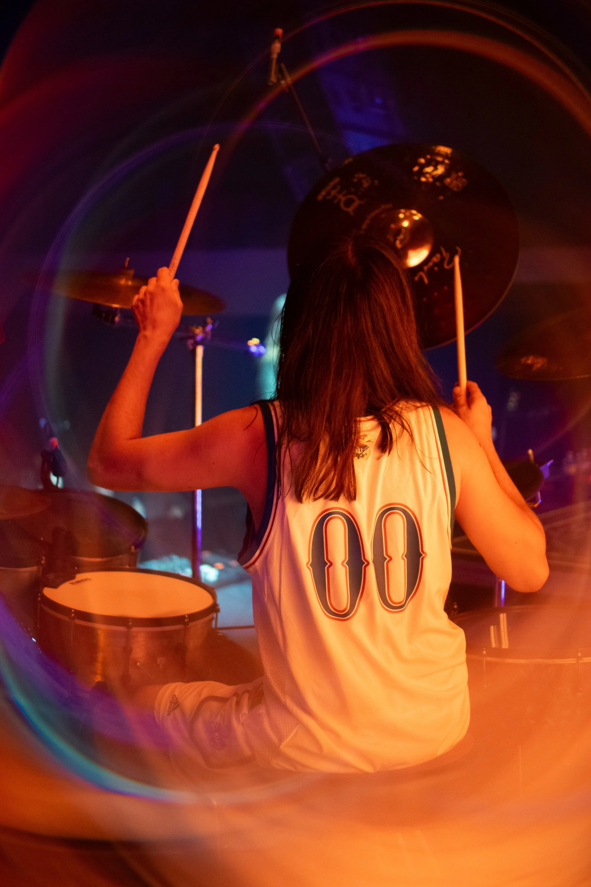 A woman with shoulder-length hair playing drums on stage under colorful lights, wearing a white sports jersey with the number 00 on the back.