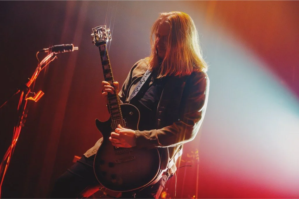 A musician with long blond hair playing an electric guitar on stage, illuminated by colorful stage lighting.