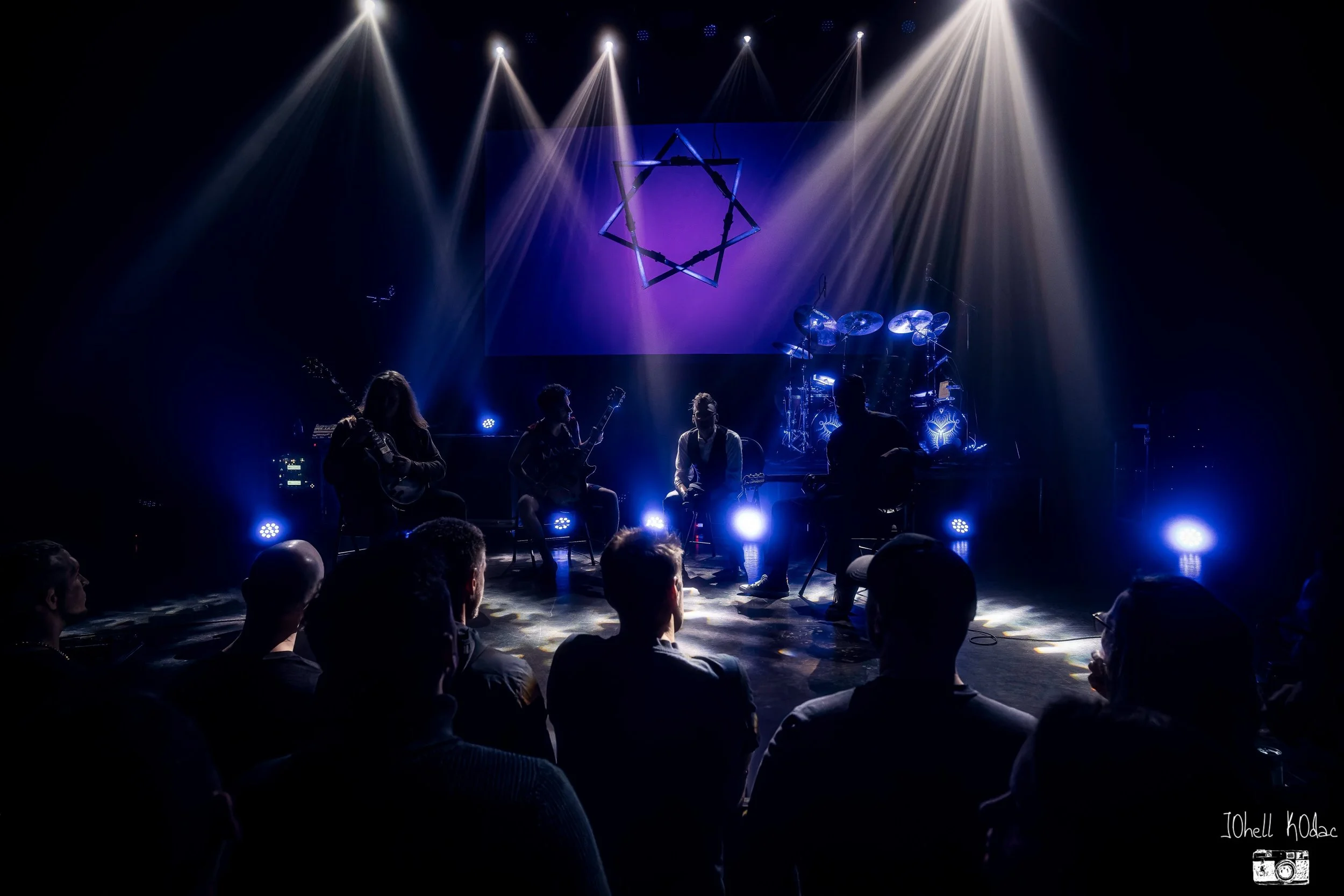 A band performing on stage with bright spotlights shining down, featuring four musicians with instruments, audience members facing the stage, and a geometric light installation hanging above.