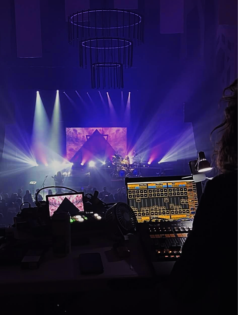 Concert stage with colorful lights and a video screen, viewed from the back of the stage with a lighting and sound technician at the mixing console in the foreground.