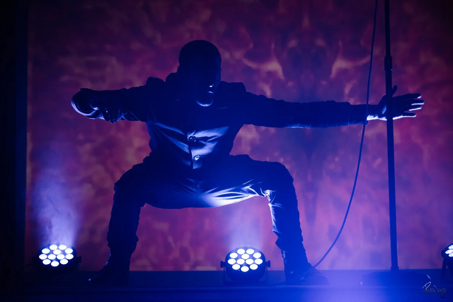 Silhouette of a man in a crouching pose on stage, illuminated by purple and blue stage lights, with a backdrop of swirling colors and holding a vertical pole with one hand.