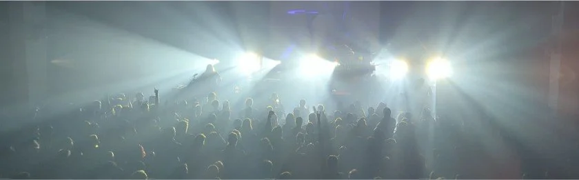 Crowd of people at a concert or event with bright stage lights shining from the front.