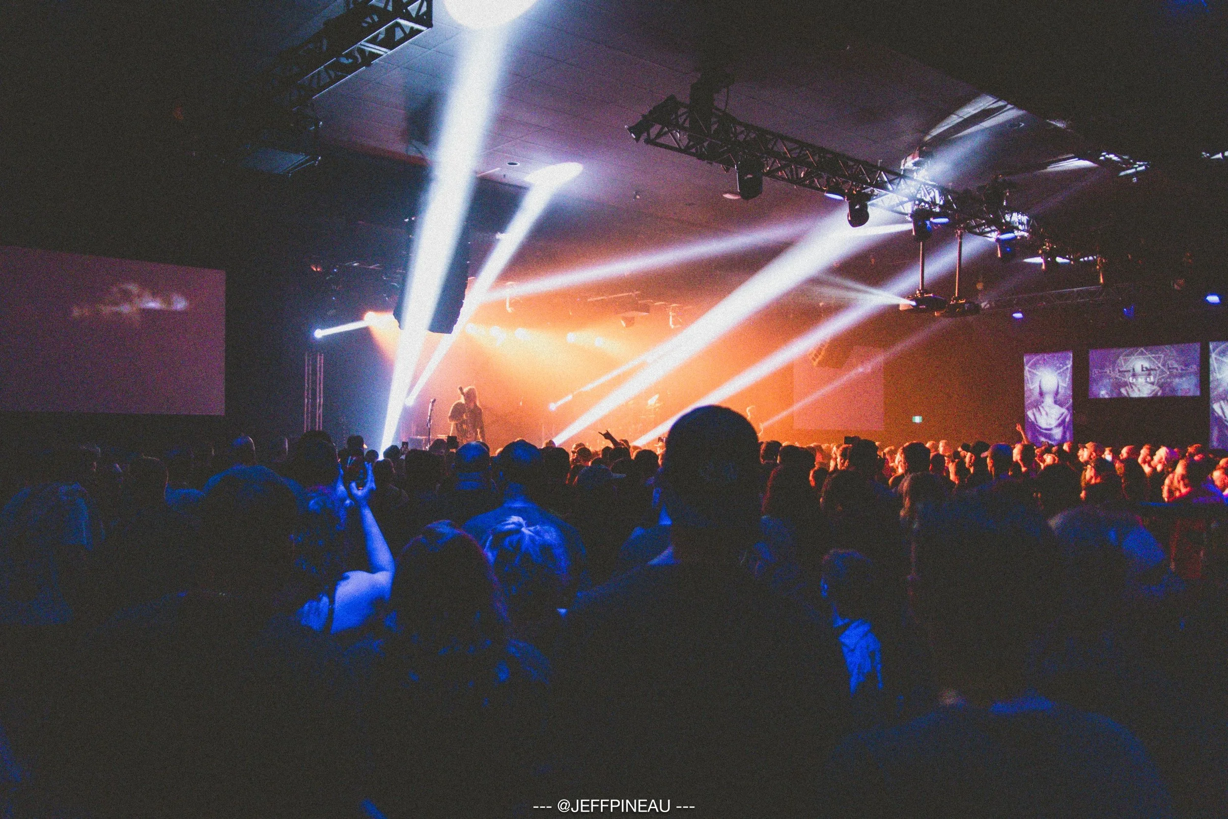 An indoor concert scene with a large crowd watching a performer on stage, illuminated by bright white and orange stage lights, with some videos or images displayed on screens on the sides of the stage.