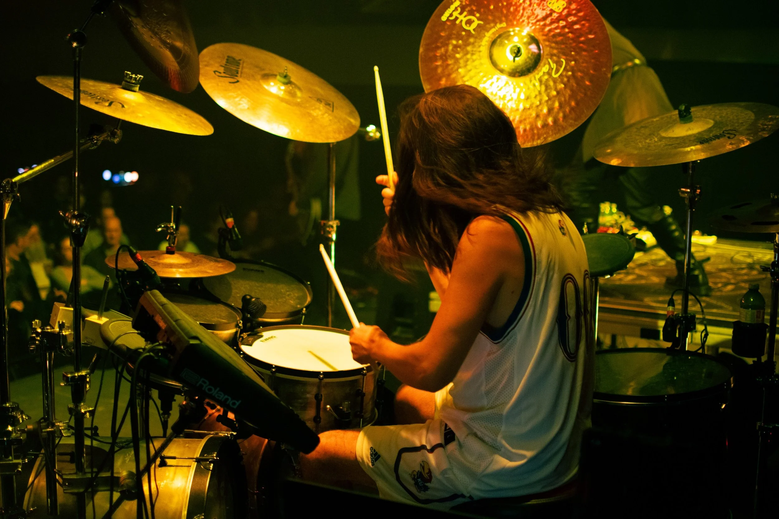 A drummer playing electronic drum set during a concert or performance, with an audience visible in the background.