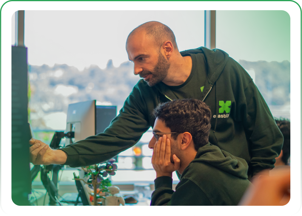 Two men working at a computer in an office, with one man explaining something to the other.