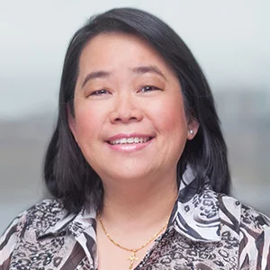 A woman smiling with black hair, wearing a floral patterned blouse and a gold necklace, standing outdoors with a blurred background.