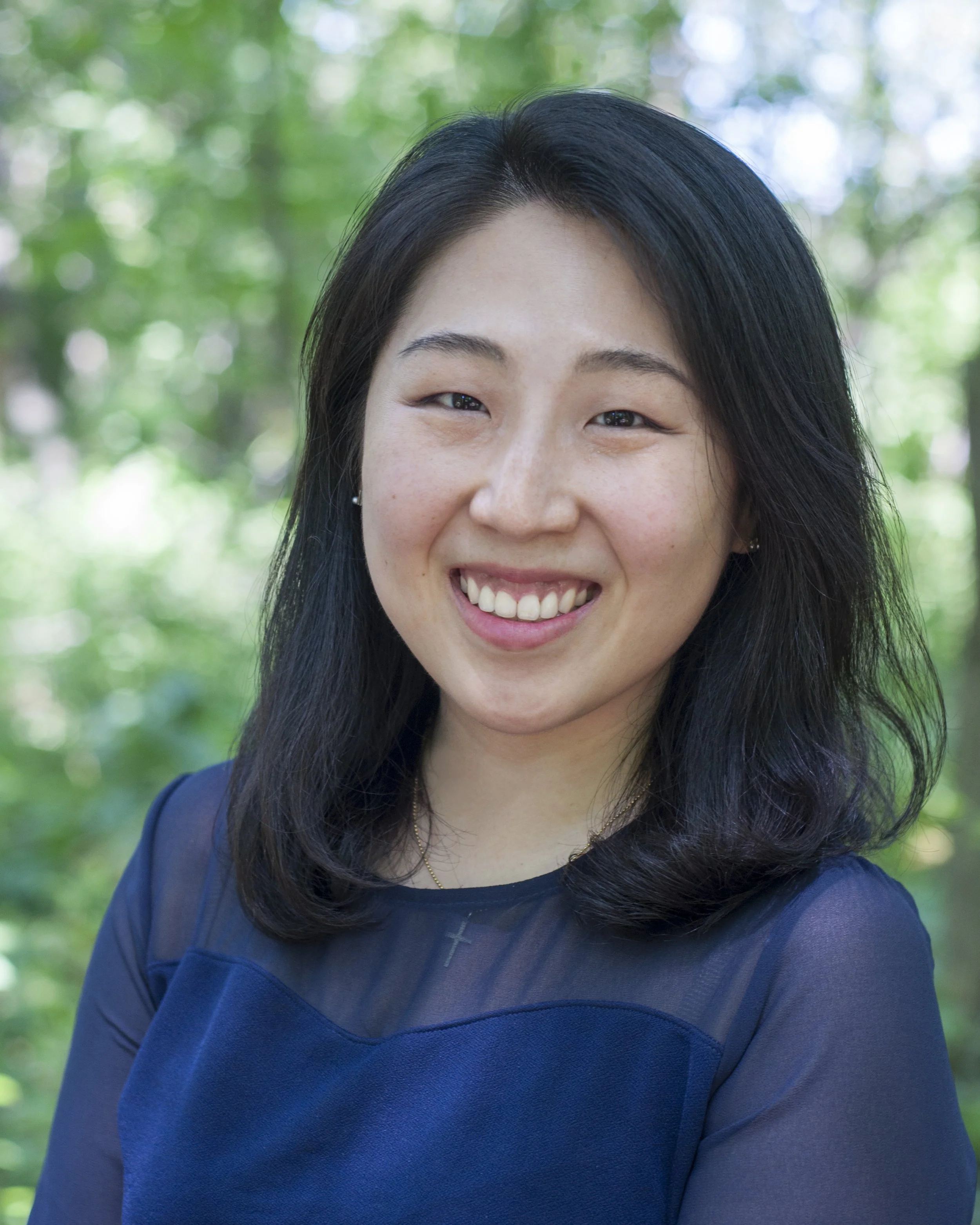 Close-up of a smiling woman with long black hair, wearing a navy blue top and small earrings, outdoors with blurred green foliage in the background.