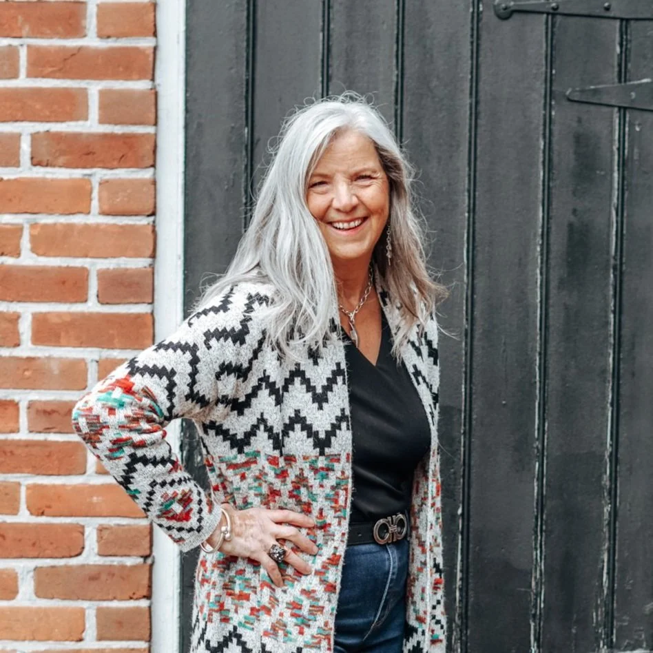 A smiling woman with long gray hair standing in front of a brick wall and a black wooden door, wearing a patterned cardigan, black top, and jeans.