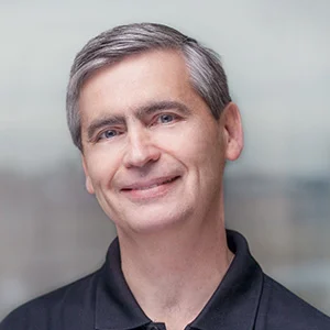 A middle-aged man with gray hair smiling, wearing a dark collared shirt, standing outdoors with a blurred background.