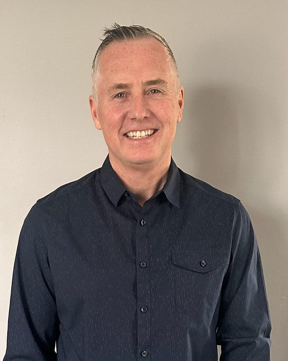 A smiling man with short gray hair wearing a dark button-up shirt, standing against a plain wall.