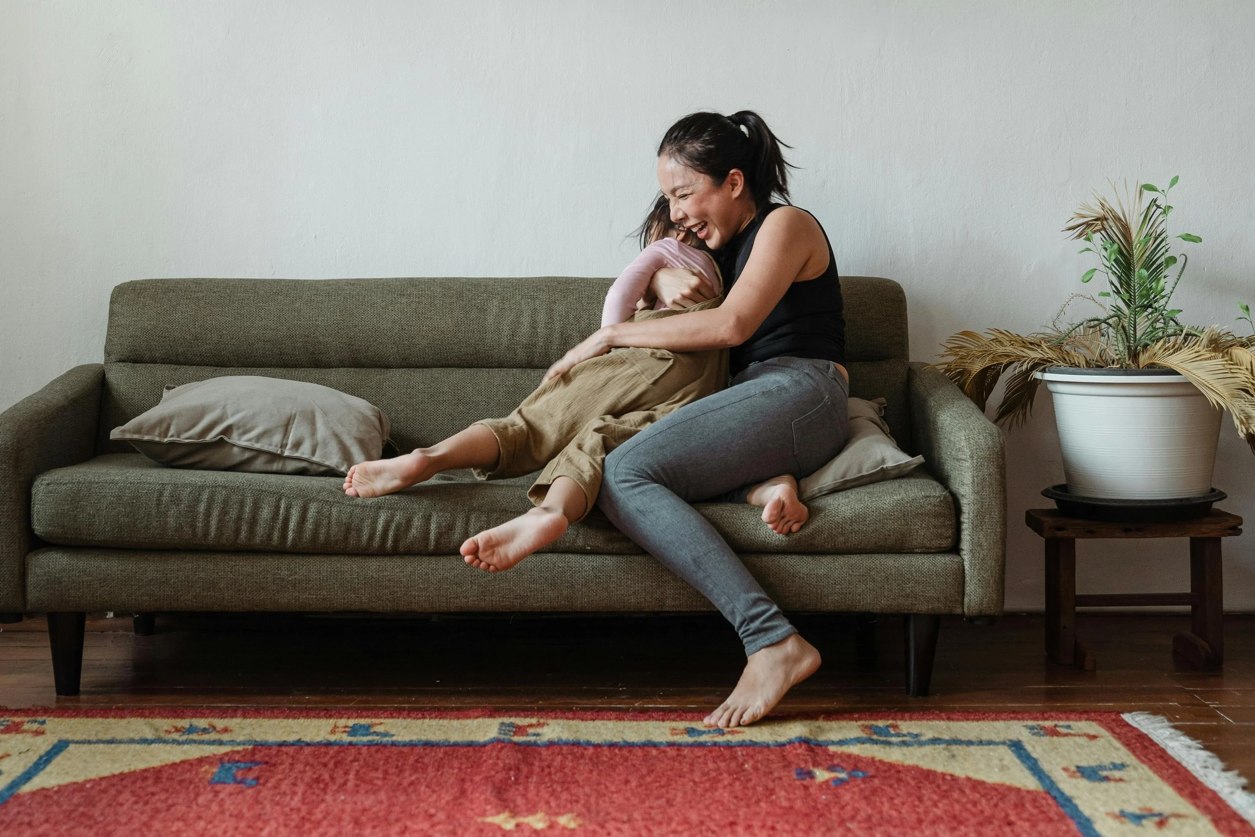 A woman and a young girl sitting on a green couch, hugging and smiling. The woman has dark hair tied back, is wearing a black tank top and gray pants. The girl has light skin, is wearing beige pants and a pink top. There are pillows on the couch, a large potted plant on a side table, a colorful rug on the floor, and a plain white wall in the background.