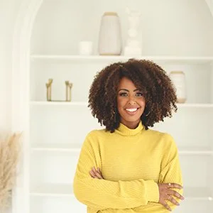 Smiling woman with curly hair wearing a yellow sweater standing in a bright, minimalist room with white shelves.