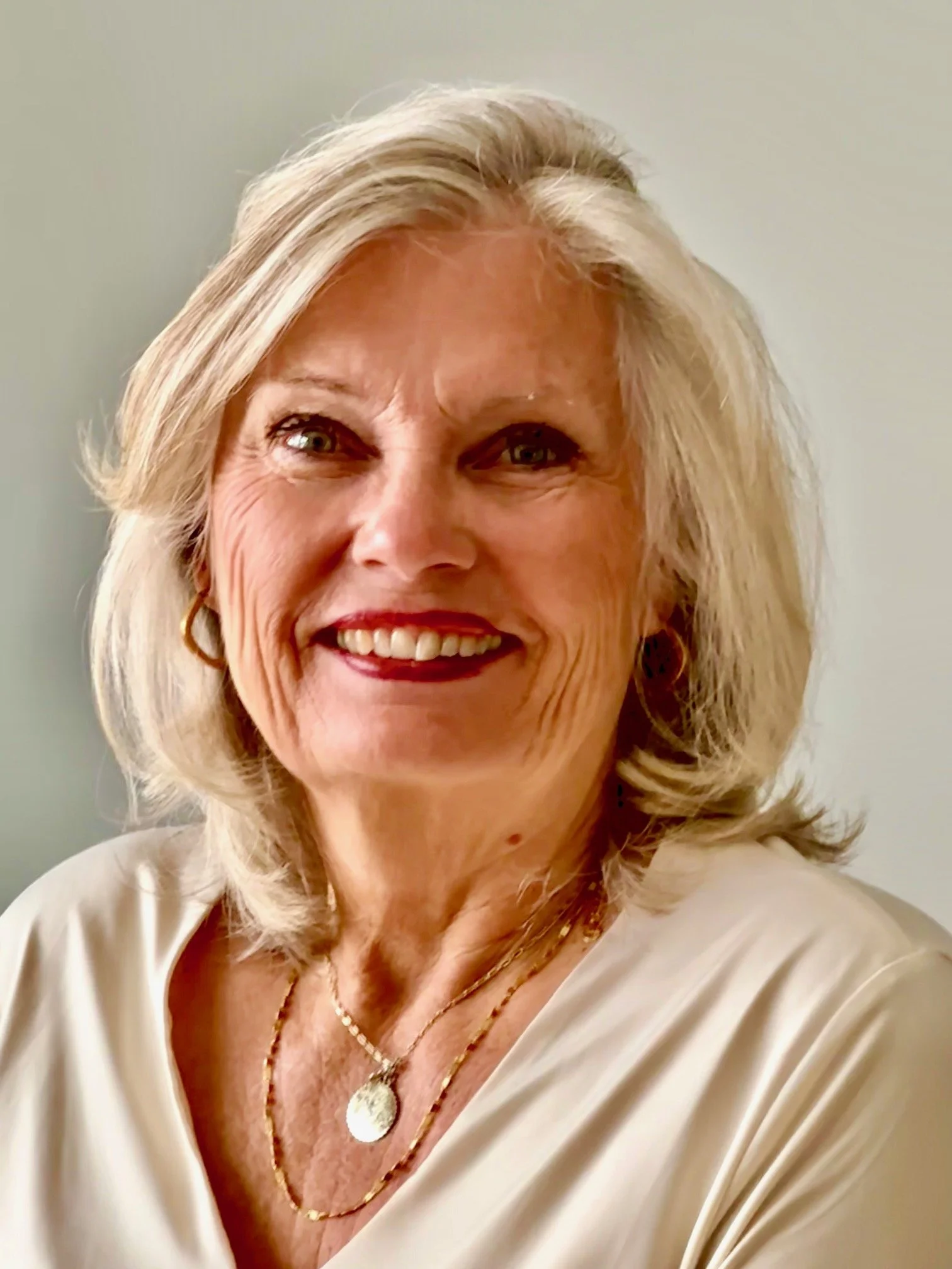 Close-up of an elderly woman with shoulder-length gray hair, smiling, wearing a cream-colored top and layered necklaces.
