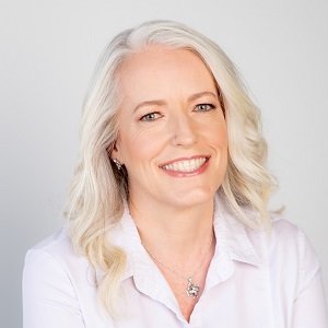 Portrait of a smiling middle-aged woman with shoulder-length white hair, wearing a white top and jewelry, against a plain light background.