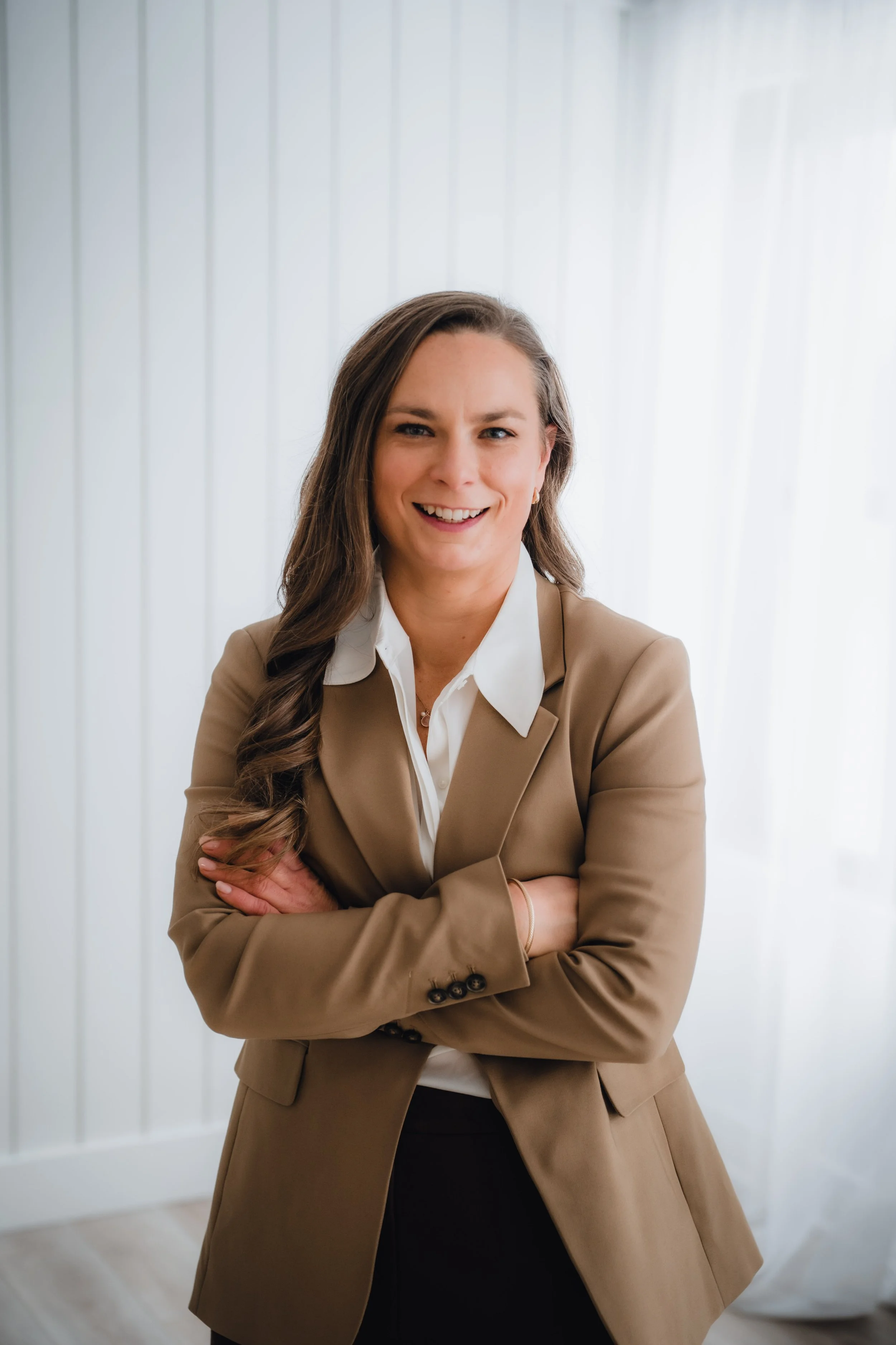 A woman with long brown hair in a loose braid, wearing a beige blazer over a white shirt, smiling with arms crossed, standing in front of a white paneled wall and sheer curtains.