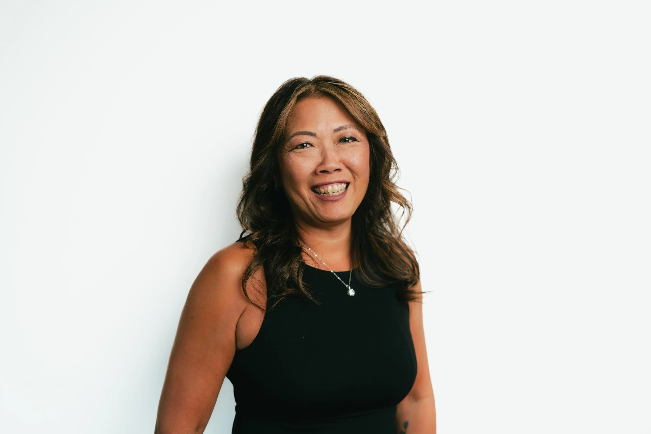 A smiling woman with long wavy hair wearing a black sleeveless top and a silver necklace, standing against a plain white background.