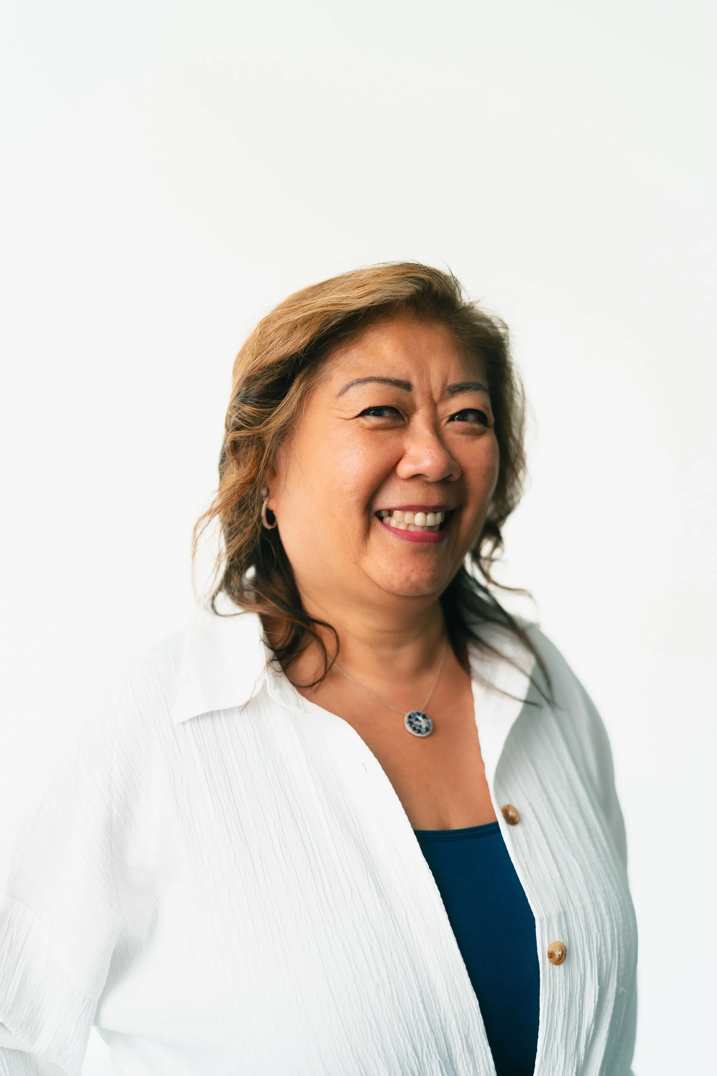A woman smiling, wearing a white textured blazer, a blue top, a necklace with a round pendant, and earrings, against a plain white background.