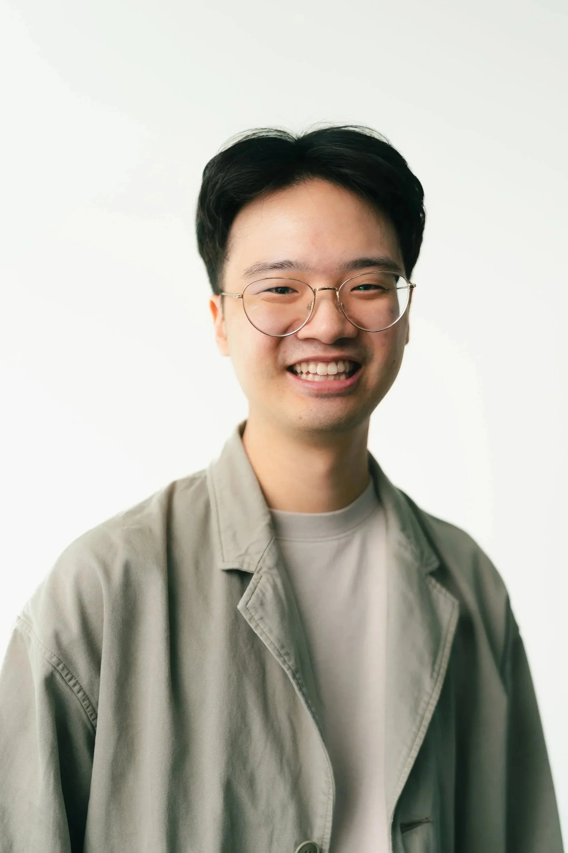 Young man with short dark hair and glasses smiling, wearing a light-colored jacket over a white shirt, against a plain white background.
