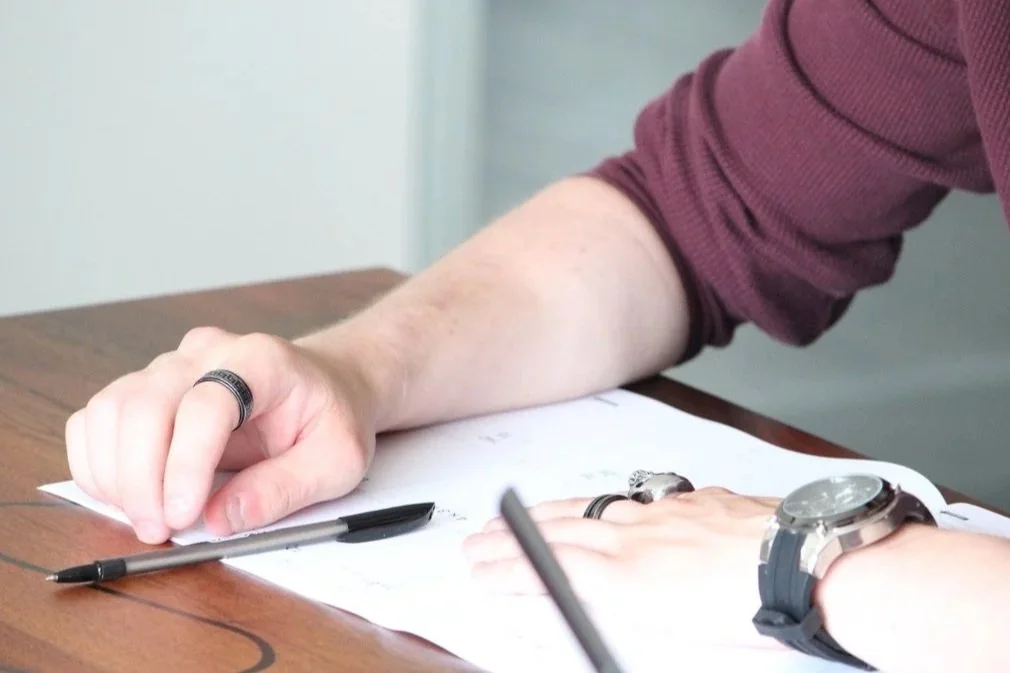 Person wearing a maroon long-sleeve shirt writing on paper with a black pen, wearing a watch and rings, on a wooden table.