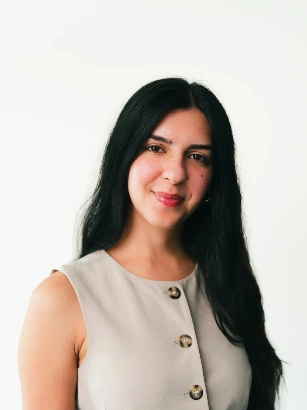 A woman with long black hair, wearing a sleeveless beige top with large metallic buttons, smiling slightly against a plain white background.