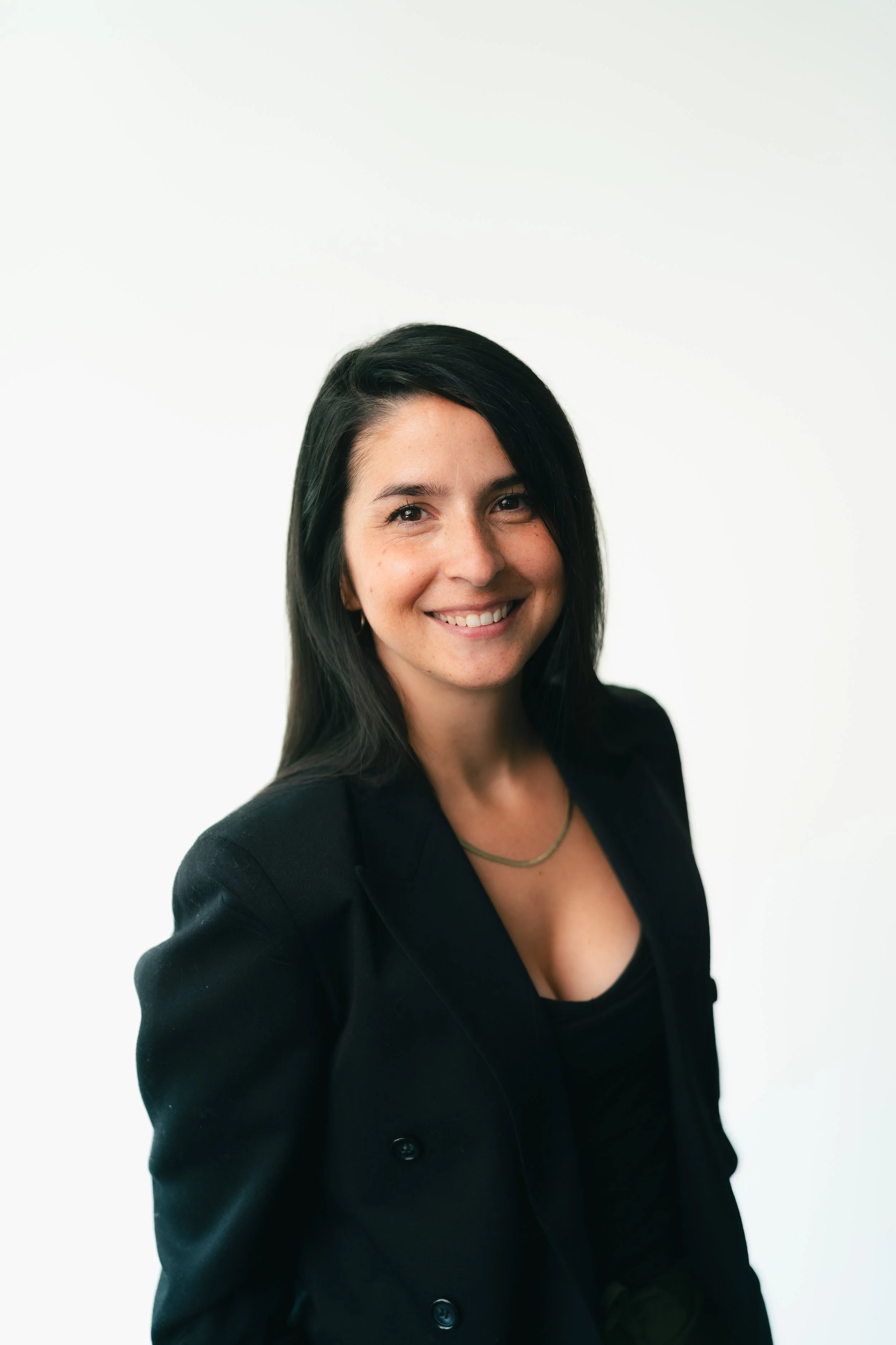 A woman with black hair, wearing a black blazer over a black top, smiling, against a plain white background.