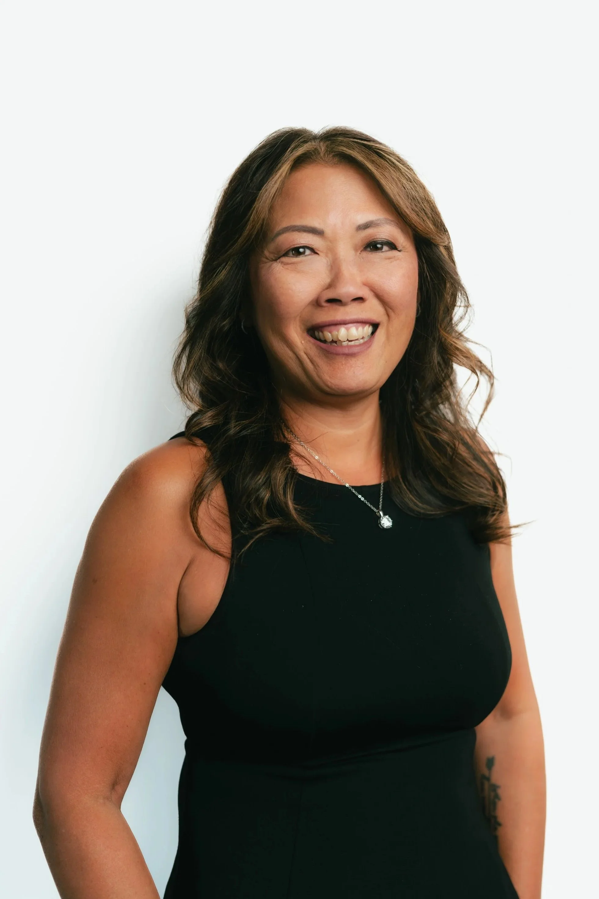 A woman with dark wavy hair, wearing a black sleeveless top, smiling, with a necklace and tattoos on her right arm, standing against a plain white background.