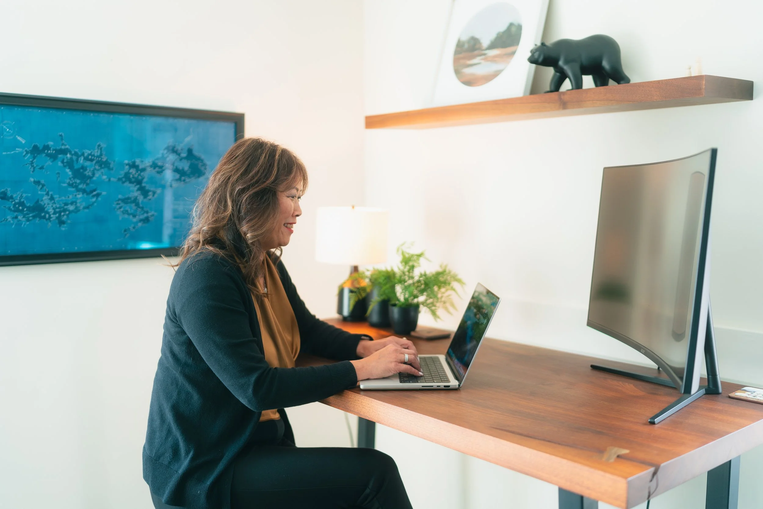 A woman with brown hair sitting at a wooden desk working on a laptop, with a large desktop monitor and a potted plant. There is a framed picture of a landscape and a black bear figurine on a wooden shelf behind her.