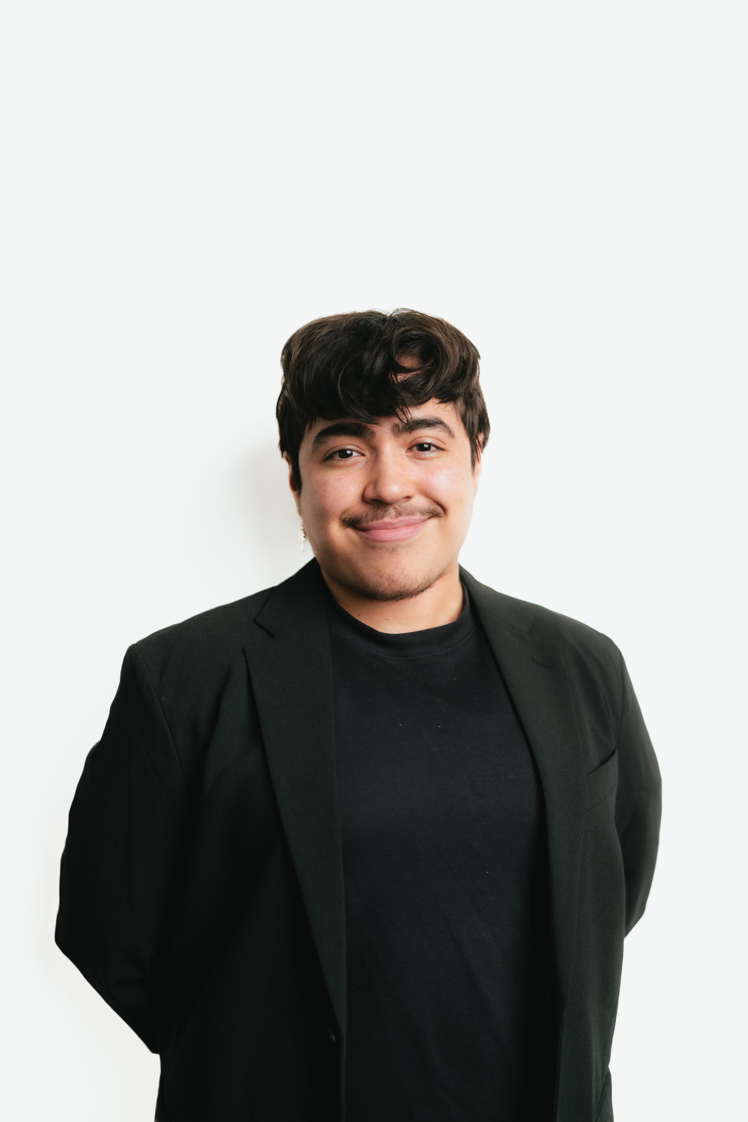 A young man with dark hair, mustache, and goatee, smiling slightly, wearing a black blazer and t-shirt, standing against a plain white background.