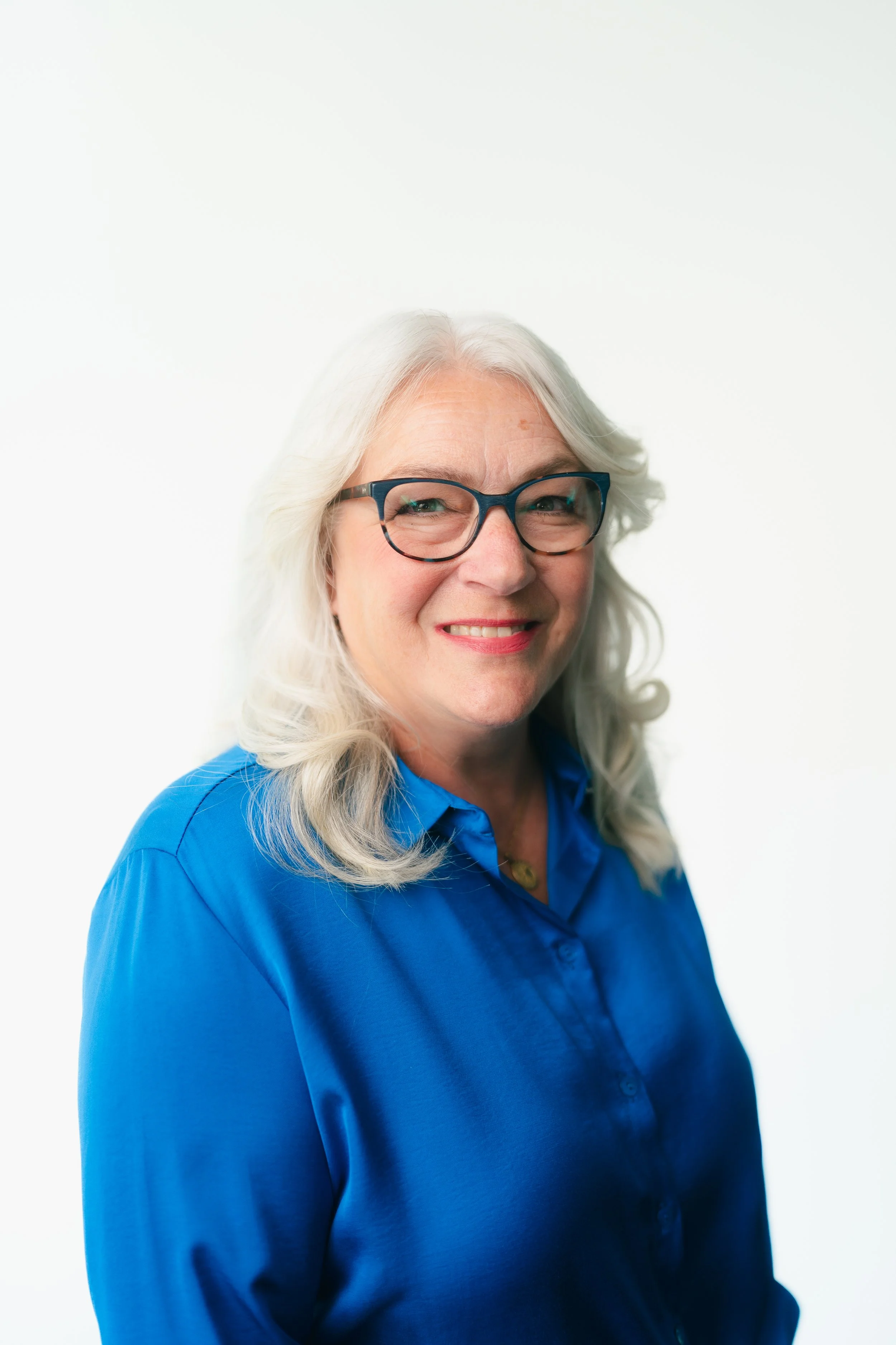 A smiling elderly woman with white hair, glasses, and fair skin, wearing a blue button-up shirt, against a plain white background.