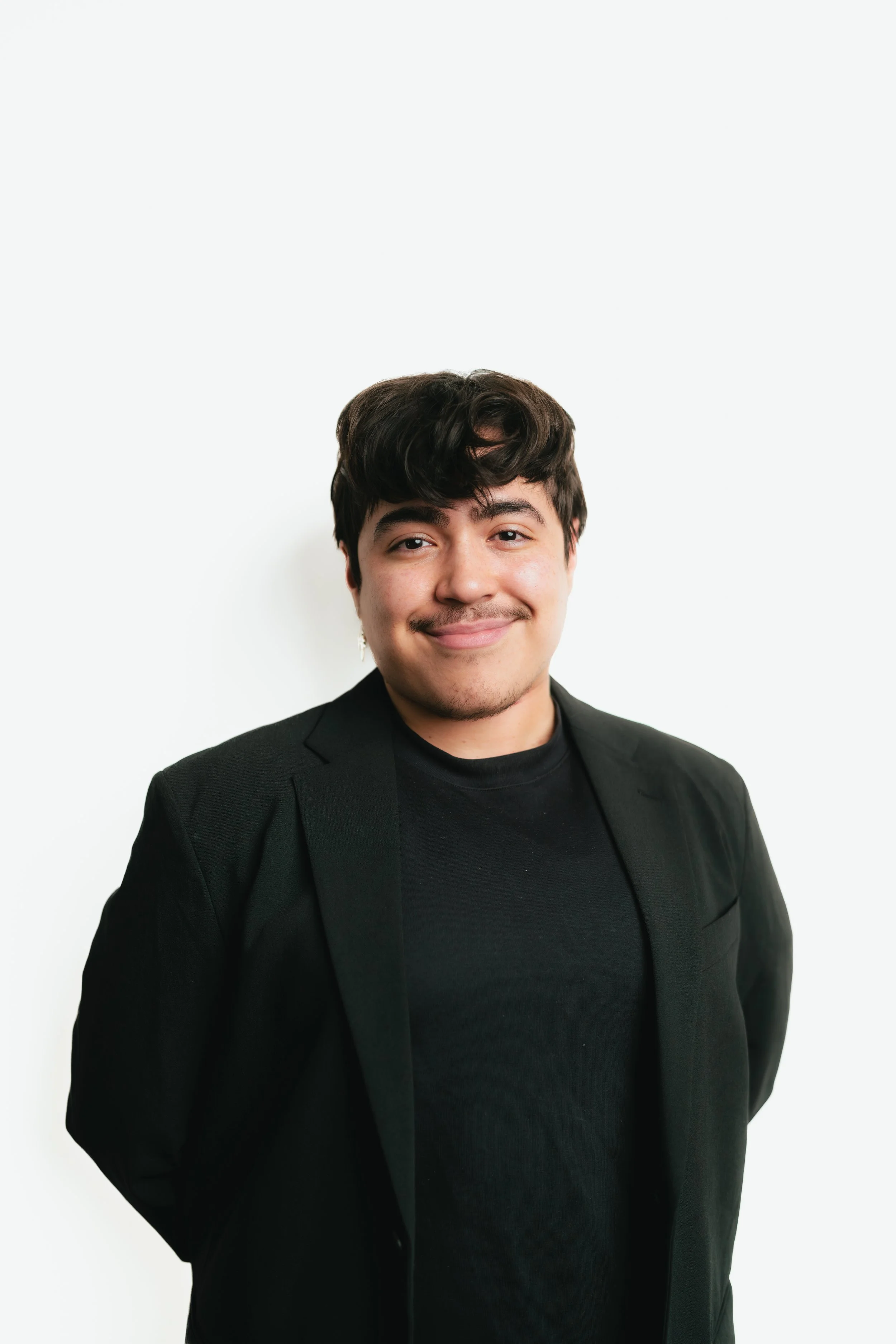 Portrait of a young man with dark hair, mustache, and beard, wearing a black blazer over a black shirt, smiling against a plain white background.