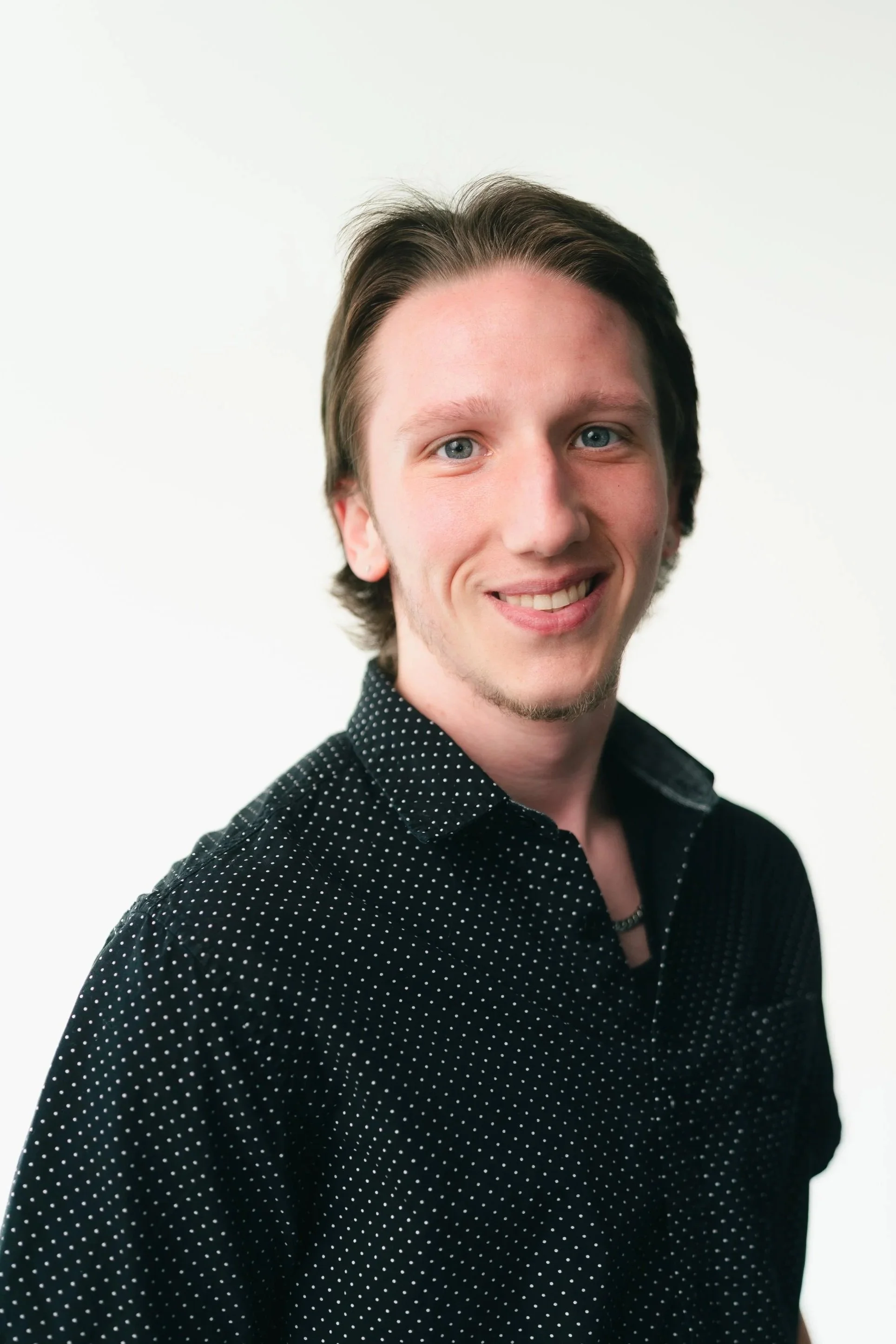 Headshot of a young man with light skin, straight brown hair, blue eyes, and a friendly smile, wearing a black polka dot shirt against a white background.