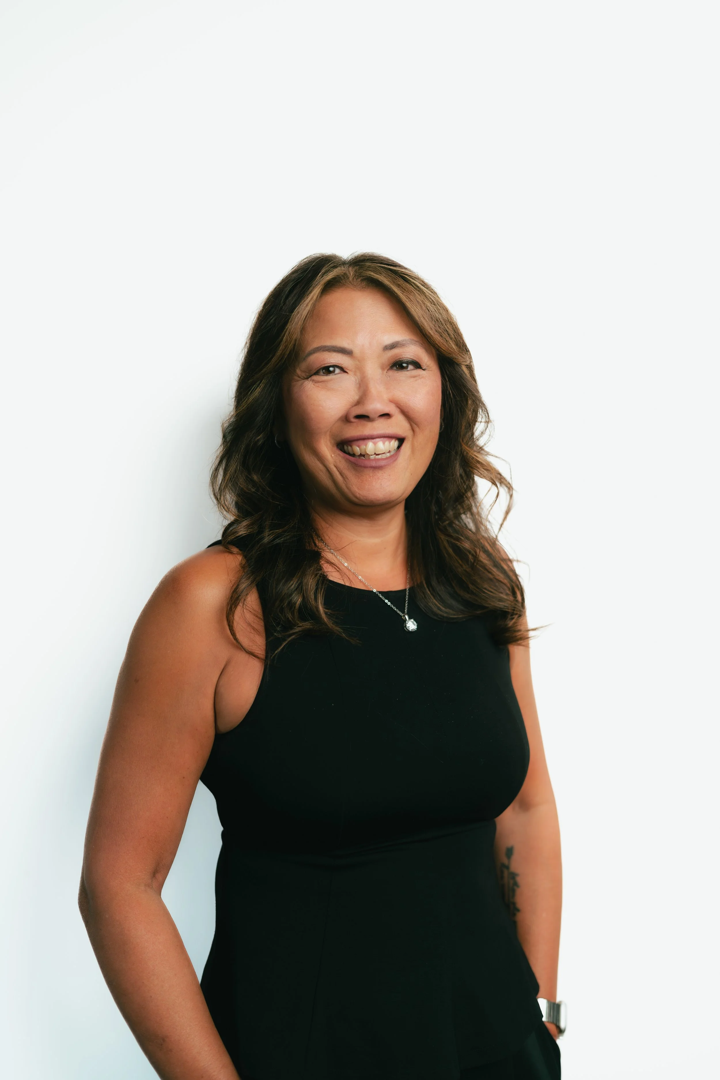 A woman with shoulder-length wavy brown hair, wearing a black sleeveless top, smiling at the camera, against a plain white background.
